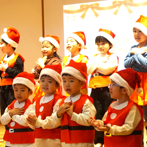 サンタクロースの帽子と赤いベストを着て、舞台上で歌っている子供たち