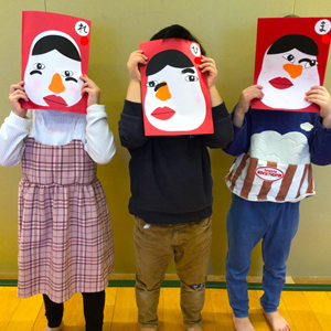 大きな顔の絵が描かれた紙を顔の前に掲げる3人の子供たち