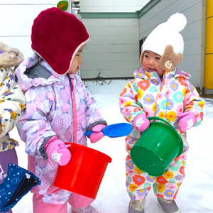雪の中でカラフルな雪遊び用ウェアを着て、シャベルとバケツで遊ぶ2人の幼児