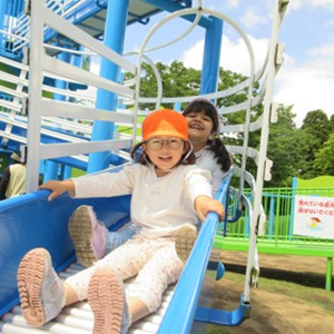 2人子供たちが、屋外の青い滑り台で楽しそうに遊んでいる。手前の子供はオレンジ色の帽子をかぶり、後ろの子供は笑顔で滑り台から顔を出している
