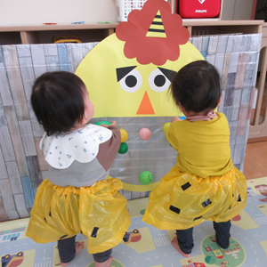 明るい色の衣装を着た幼児2人が、鬼の顔の的当てで遊んでいる