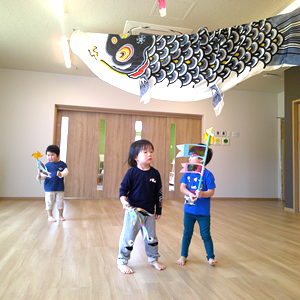 屋内の広い空間に、大きな鯉のぼりが飾られている。3人の幼児がそれぞれ小旗のようなものを持って立っている。