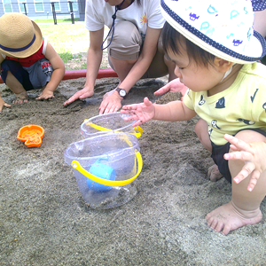 屋外の砂場で、複数の幼児と大人が砂遊びをしている。手前の幼児は黄色いバケツに手を伸ばし、奥の幼児はシャベルで砂を掘っている。