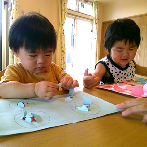屋内で、2人の幼児がテーブルで工作をしている。手前の幼児は紙に貼られた小さな紙片を触っており、もう一人の幼児も紙を見つめている。