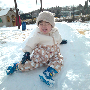 雪が積もった屋外で、ベージュの雪遊び用ウェアを着た幼児が、雪の小さな山に座って満面の笑みを浮かべている。背景には遊具や木々が見える。