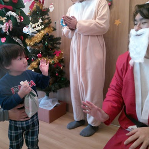 屋内で、クリスマスツリーのそばでサンタクロースの衣装を着た人と、動物の着ぐるみを着た人が立っている。一人の幼児がプレゼントらしきものを持ってサンタクロースの前に立っている。