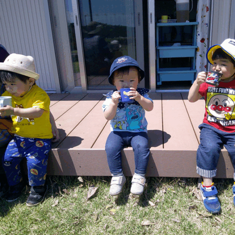 屋外のウッドデッキに座っている3人の幼児。全員がコップで飲み物を飲んでいる。手前の幼児は麦わら帽子をかぶり、中央の幼児は紺色の帽子をかぶっている。右奥の幼児は黄色い帽子をかぶっている。
