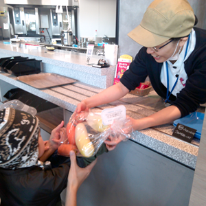 店内で、カウンター越しに女性店員らしき人物が子供に野菜の入った袋を手渡している。