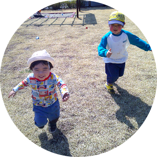 屋外の芝生の広場で、2人の幼児が楽しそうに走っている。手前の幼児は白い帽子をかぶり、もう一人の幼児は黄色い帽子をかぶっている。
