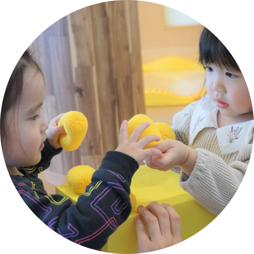 屋内で、2人の幼児が黄色いポンポン状のおもちゃで遊んでいる。一人の幼児がもう一人の幼児にポンポンを渡しているように見える。