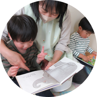 マスクを着用した保育士が、屋内で2人の幼児に絵本を読み聞かせている。一人の幼児は絵本を指差し、もう一人の幼児は隣で座っている。