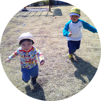 屋外の芝生の広場で、2人の幼児が楽しそうに走っている。手前の幼児は白い帽子をかぶり、もう一人の幼児は黄色い帽子をかぶっている。