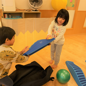 屋内の広い空間で、二人の幼児がバランスボードのような青い遊具で遊んでいる。床は木目調で、奥には緑色のボールが見える。
