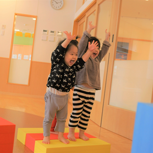 屋内の広々とした空間で、二人の幼児が積み木のようなブロックの上に立ち、両手を高く上げて楽しそうにしている。