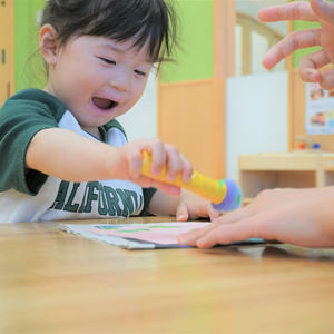屋内で、一人の幼児がテーブルに置かれた紙に向かって、黄色のペン型ツールを使い、もう一人の大人の手がそばにある。幼児は笑顔で集中しているように見える。