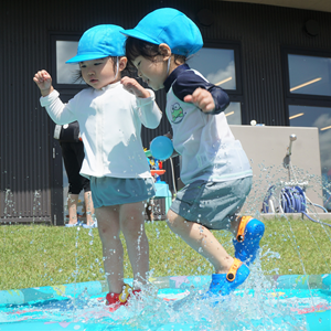 屋外で、二人の幼児が小さなプールのような水場で水遊びをしている。二人は水着と青い帽子を着用しており、水しぶきが上がっている。