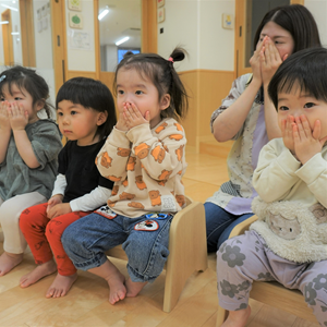 屋内で、複数の幼児と一人の先生が口に手を当てて座っている様子。