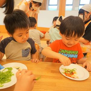 屋内で、複数の幼児がテーブルを囲んで食事や調理体験をしている様子。手前の幼児は、皿の上の食材に手を伸ばしている。