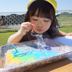 外で、黄色い帽子をかぶった女の子が、トレーに入ったカラフルな液体と、その中に浮かぶ何かを興味深そうに見つめている。背景には鯉のぼりが見える。
