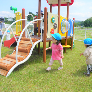 屋外の芝生のある広場で、複数の幼児が青い帽子をかぶり、色とりどりの遊具で遊んでいる。