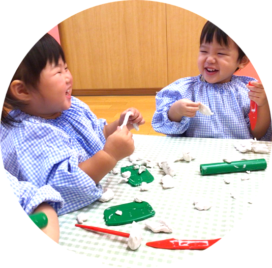 青いチェックのスモックを着て、笑顔で粘土遊びを楽しむ二人の子ども。