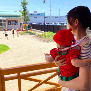 保育士に抱っこされたスイカ柄の服を着た赤ちゃんが、テラスからお兄さんお姉さんが遊ぶ園庭を指さして眺めている姿。