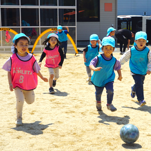 園庭でビブスを着て、一生懸命にボールを追いかけながらサッカーを楽しむ子どもたちの躍動感あふれる様子。
