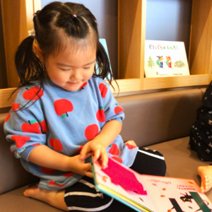 りんご柄の可愛い服を着た女の子が、ソファに座って熱心に絵本をめくっている微笑ましい一コマ。