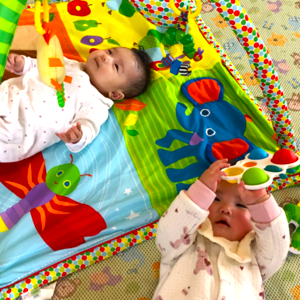 カラフルなベビージムに寝転がり、吊り下げられたおもちゃに興味津々で手を伸ばして遊ぶ二人の赤ちゃん。