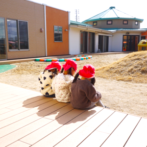 ぽかぽか陽気のウッドデッキに並んで座り、お揃いの赤い帽子をかぶって園庭を眺める三人の子どもの後ろ姿。