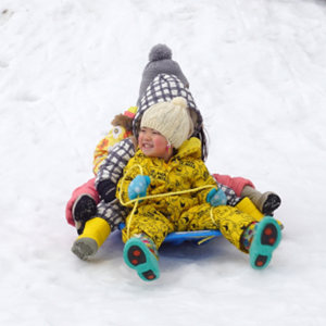 雪山で青いソリに乗り、防寒着を着て満面の笑みで滑り降りる子どもたちの楽しそうな冬の風景。