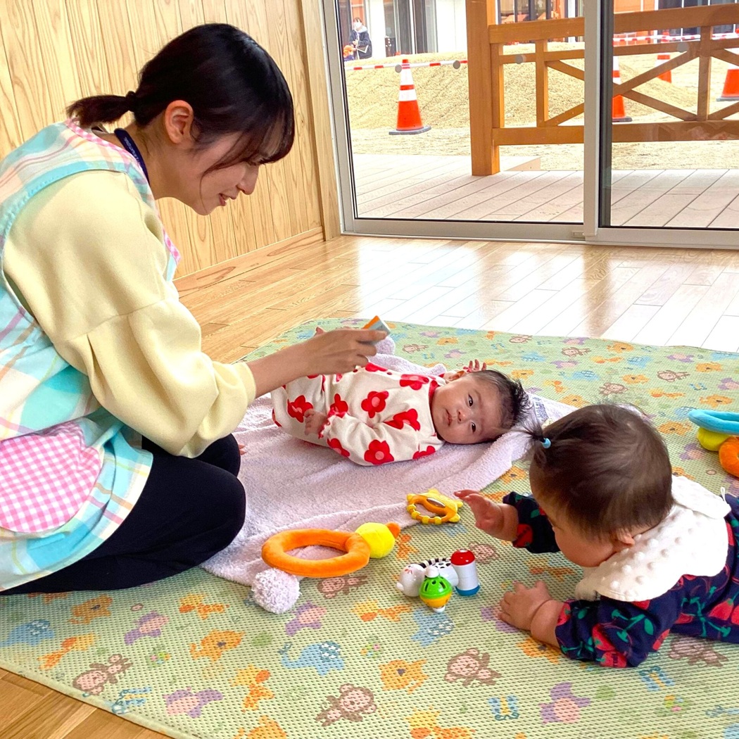室内で保育士に見守られながら、マットの上で遊ぶ二人の乳児。