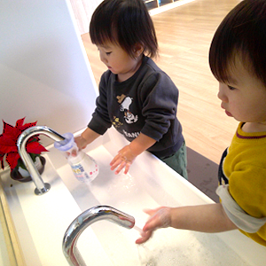 洗面台で保育士に見守られながら、石鹸を泡立てて一生懸命に手を洗っている二人の子ども。