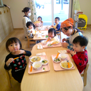 保育室でテーブルを囲み、先生と一緒に楽しそうに給食（スパゲッティなど）を食べている子どもたちの食事風景。