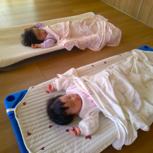 お昼寝用のコットの上で、タオルケットを掛けて心地よさそうに眠っている二人の赤ちゃんの寝顔。