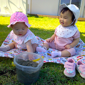 晴れた日の芝生の上で、レジャーシートを広げて座り、砂の入ったバケツを前に穏やかに過ごす二人の赤ちゃん。