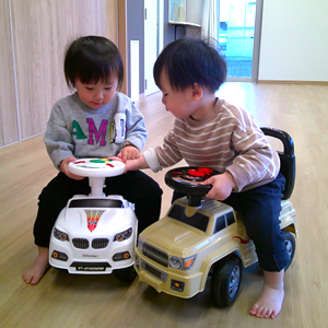 室内で白い乗用玩具とベージュの乗用玩具にまたがり、ハンドルを握って楽しそうに遊ぶ二人の子ども。