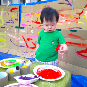 段ボールの壁一面に描かれたカラフルな絵をバックに、赤い絵の具を使って真剣に制作活動を楽しむ緑色のTシャツを着た男の子。