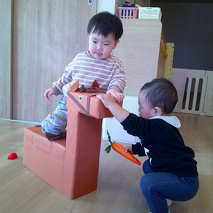 段ボールで作られた大きなお馬さんの遊具にまたがって遊ぶ子と、にんじんのおもちゃを持って寄り添う子の微笑ましい光景。
