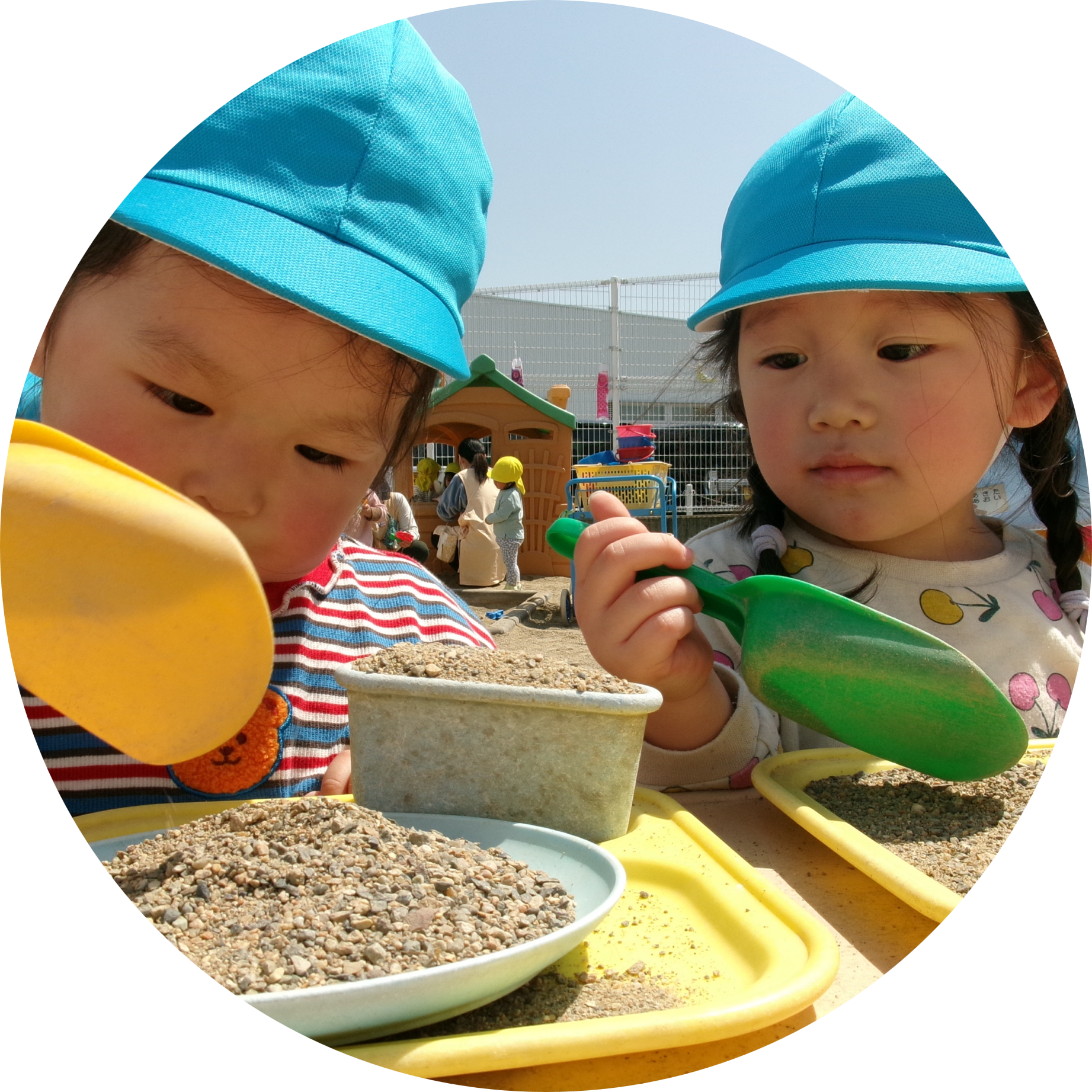 水色のカラー帽子をかぶり、真剣な表情でお皿や容器に砂を盛り付けて、お料理ごっこを楽しむ二人の子どもの様子。