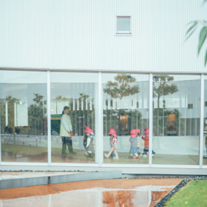 大きなガラス窓越しに、赤い帽子をかぶった子どもたちが先生に連れられて、一列になってお散歩している様子。