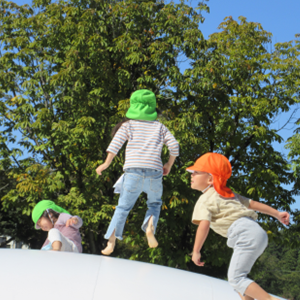 大きな木々が広がる公園で、白い雲のような大型トランポリン（ふわふわドーム）の上を、元気にジャンプして遊ぶ子どもたちの後ろ姿。