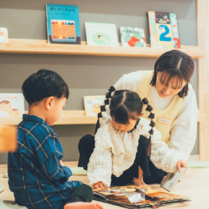 たくさんの絵本が並ぶ図書コーナーで、保育士と一緒に床に座り、一冊の本を夢中で読み進めている子どもたちの穏やかな時間。