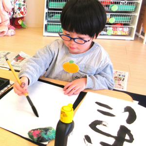 真剣な表情で筆を運び、大きな紙に力強い文字を書いている、眼鏡をかけた男の子の書道体験の様子。