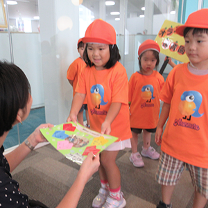 お揃いのオレンジ色のTシャツと帽子をかぶった子どもたちが、手作りのメッセージカードを優しく手渡している交流の場面。