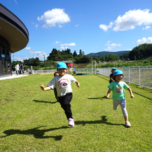 青空の下、広々とした芝生の園庭を、満面の笑みでこちらに向かって元気いっぱいに走ってくる子どもたち。