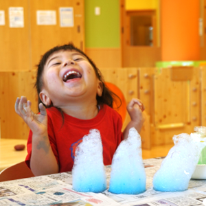 室内で大きな青い泡の塊を前にして、口を大きく開けて大笑いしながら感触を楽しんでいる女の子。