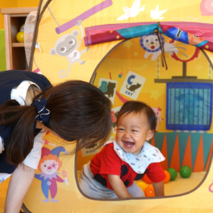 サーカス風の可愛い室内テントの中から、保育士に向かってにっこりとはじけるような笑顔を見せる赤ちゃん。