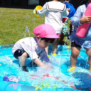 夏の陽射しのなか、ビニールプールで噴き出す水に触れて、涼しげに水遊びを満喫する子どもたちの様子。