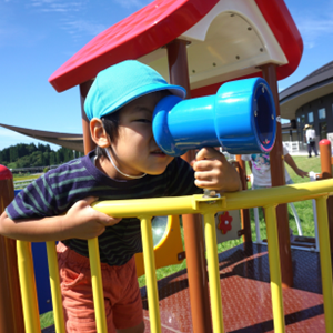 屋外遊具の上で、青い望遠鏡を真剣に覗き込み、遠くの景色を一生懸命探している男の子。